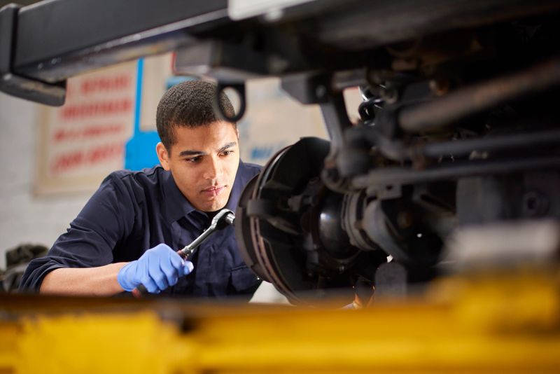 A young mechanic is working on a car in a garage repair shop. He is wearing blue overalls.  He is fixing the brake discs on the front driver's side of the vehicle.