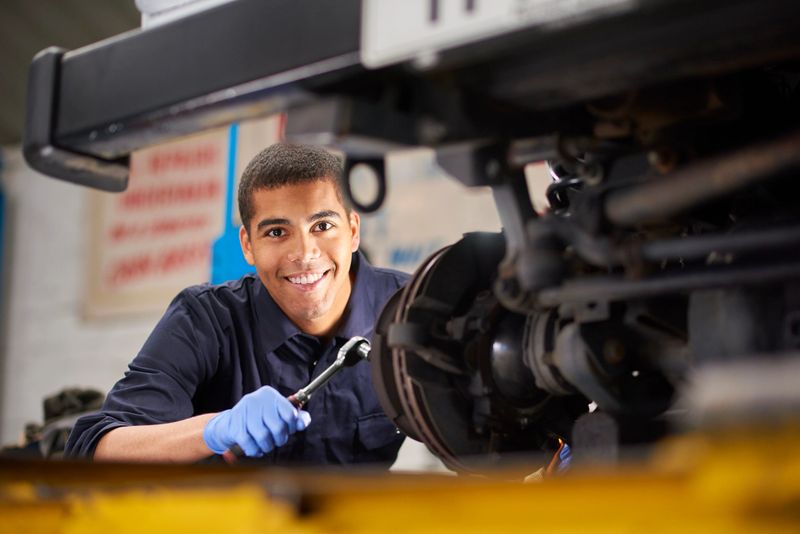 A young mechanic is smiling to camera as he works on a car in a garage repair shop. He is wearing blue overalls.  He is fixing the brake discs on the front driver's side of the vehicle.