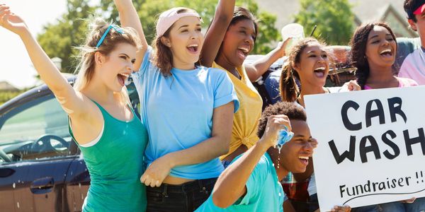 Group of young people excitedly promoting a car wash fundraiser outdoors.