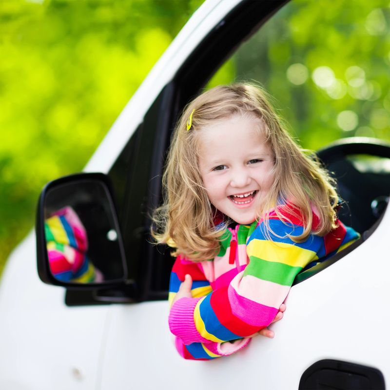 Little girl with funny pigtails watching out of car window sitting on front driver seat during a break on a family vacation road trip on summer day. Child in white minivan. Traveling by car with kids.