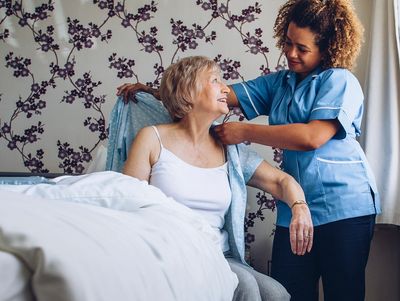 Caregiver helping elderly woman put on a robe in a cozy bedroom.
