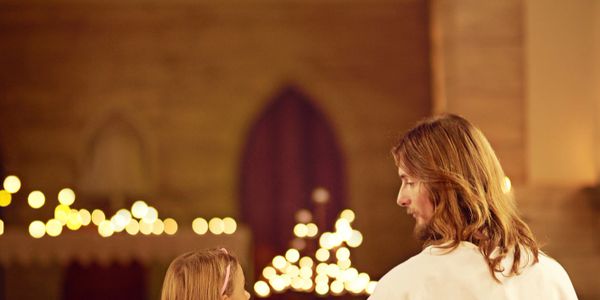 A man dressed as Jesus talks with a young girl in a church pew.