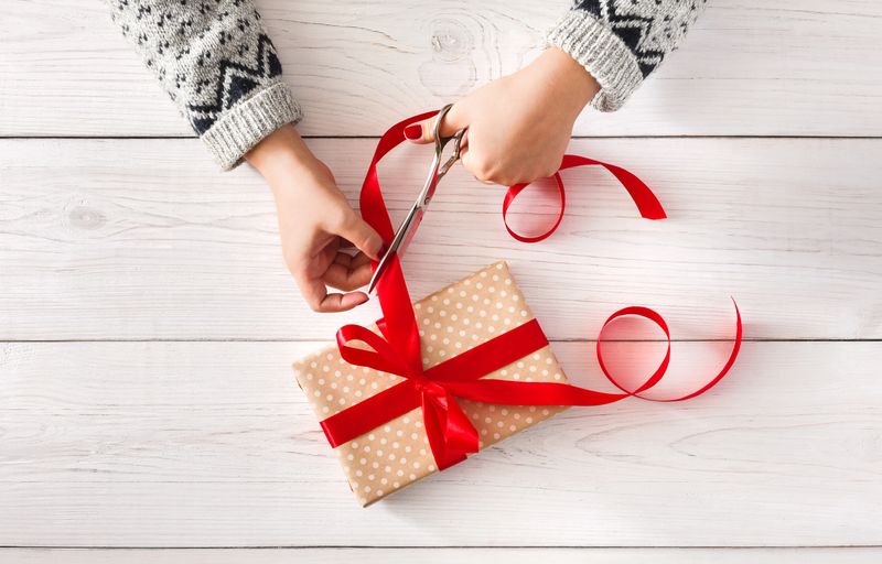 Woman is wrapping christmas or other holiday handmade present in paper with red ribbon. Making bow at present box, decoration of gift. Hands on white wooden table, top view.