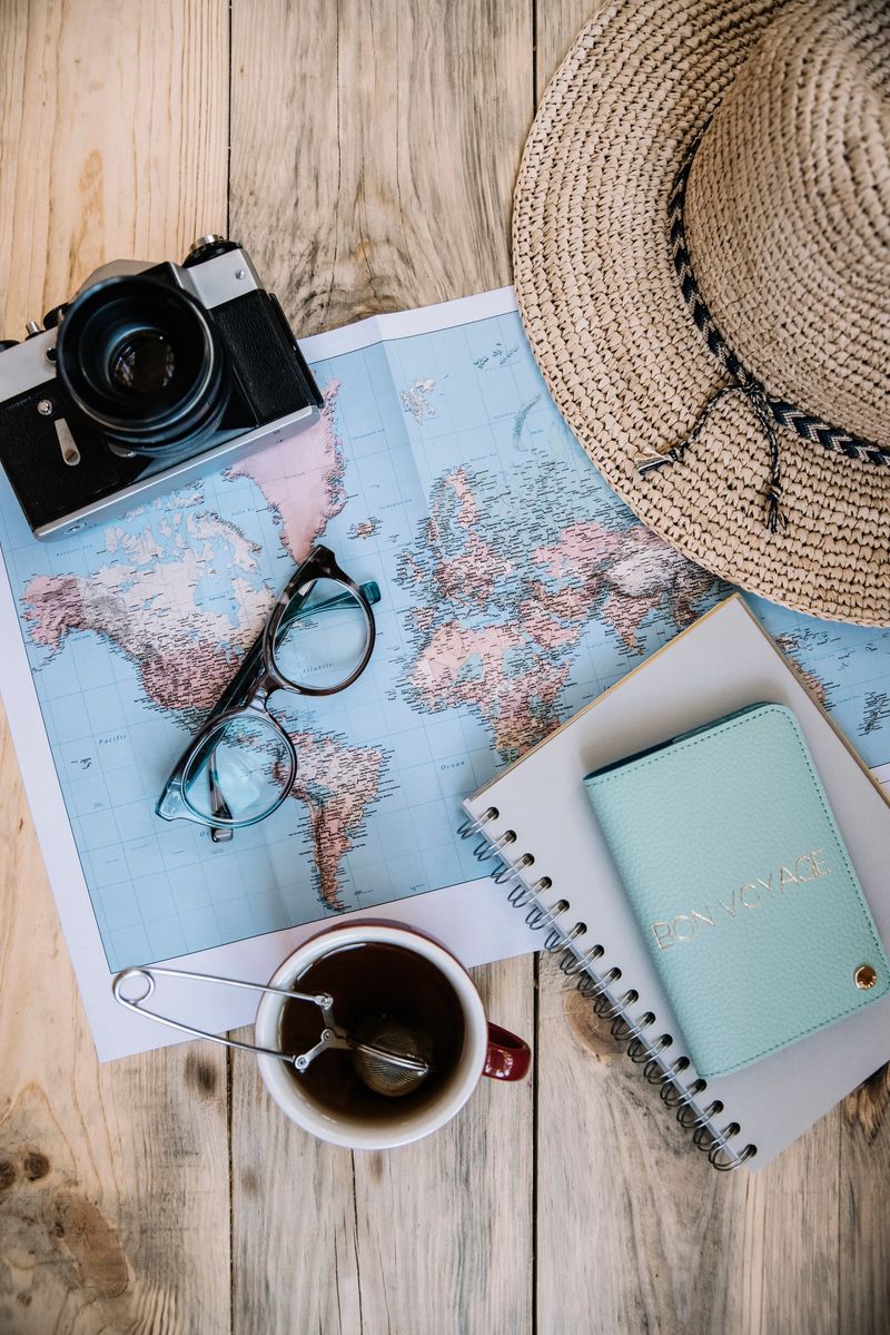 Travel preparations essentials. World map, cup of tea, vintage film camera, hat, passport, notebook and stylish glasses on the old rustic wooden table background, flat lay