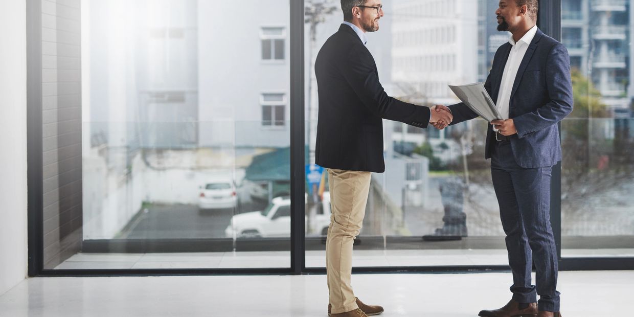 Two businessmen shake hands in a modern office with large windows.