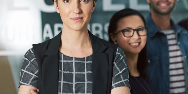 Confident businesswoman standing with crossed arms, colleagues smiling in background.