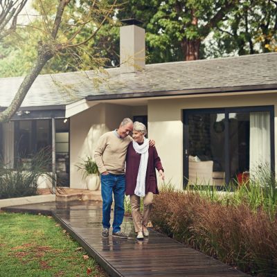 Elderly couple walking arm in arm on a wooden path outside their home.