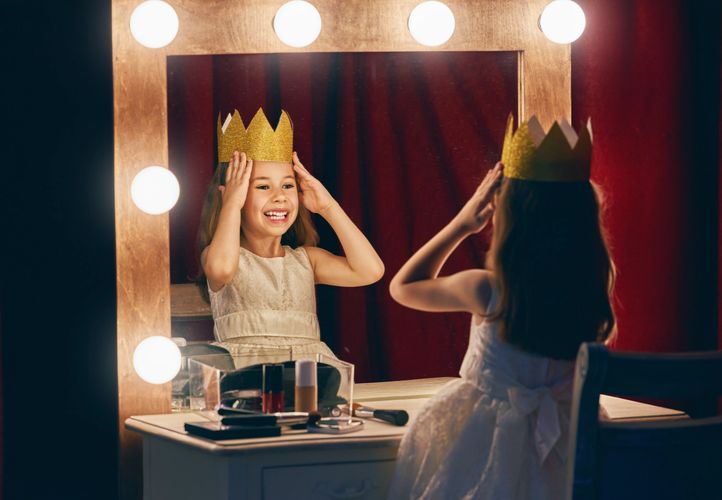 Little girl wearing a golden crown, smiling at herself in a makeup mirror.