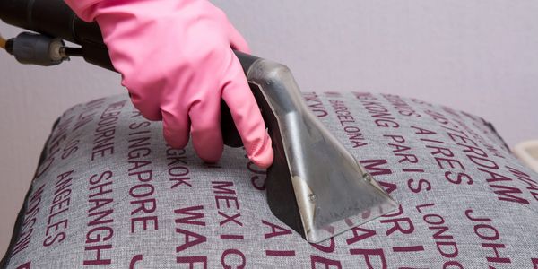 Hand with pink glove cleaning a fabric surface with a steam cleaner.