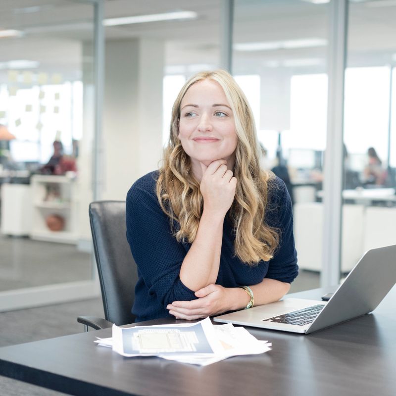 Female office worker looking away with hand on chin, woman using laptop in modern office, smiling
