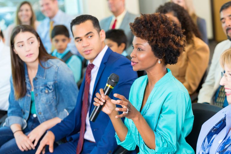 Beautiful  mature African American woman gestures while asking a political candidate a question during a town hall meeting. A diverse group of people are sitting with her in the audience.