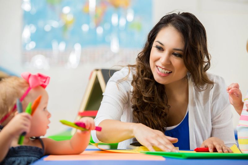 Pretty Hispanic female daycare teacher sits at table and helps cute blonde toddler girl with art project. Little toddler girl holds crayons and the teacher smiles and points to coloring page.