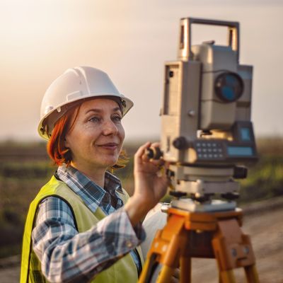 A female surveyor wearing a hard hat operates a theodolite outdoors at sunset.