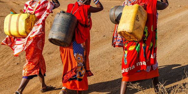 Three women in colorful traditional dresses carrying containers on their backs.