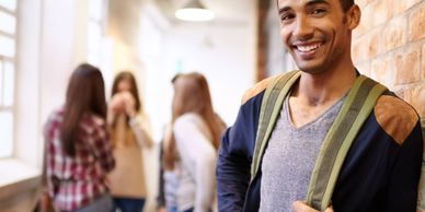 Smiling young man with backpack in school hallway, friends chatting in background.