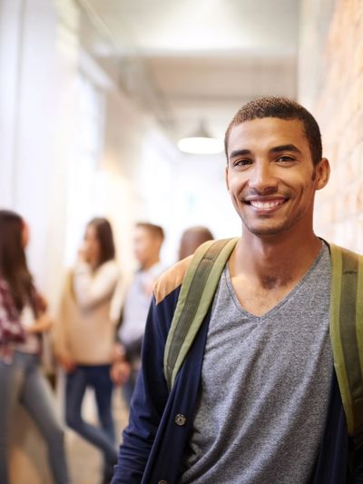 Smiling young man with a backpack leaning against a brick wall in a school hallway.
