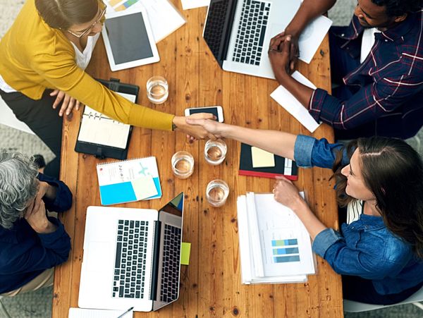 Two colleagues shake hands over a wooden table during a meeting with laptops and documents.