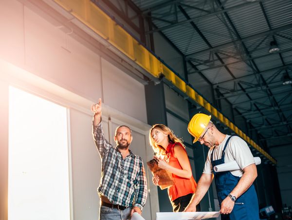 Three professionals discussing plans inside a warehouse or industrial space.