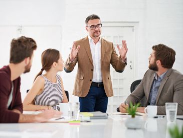 A man in a brown blazer leads a business meeting with attentive colleagues.