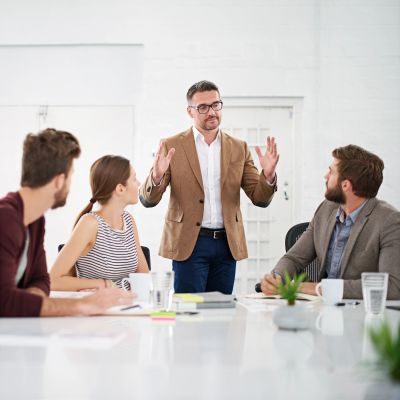 Man in brown blazer speaking to colleagues in a meeting.