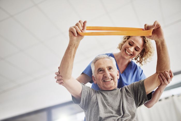 Senior man exercising with resistance band, assisted by a smiling caregiver.