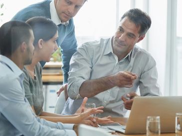 A group of four professionals engaged in a discussion around a laptop in a bright office.
