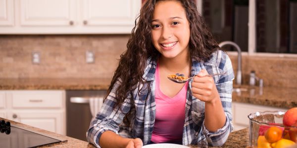 A smiling girl eating cereal with milk in a kitchen.