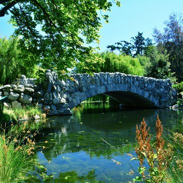 Stone arch bridge over a calm pond surrounded by lush greenery.