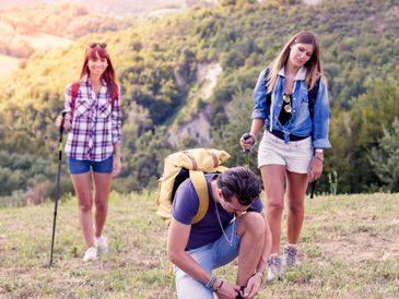 Friends hiking outdoors with one tying shoelaces on a grassy hill.