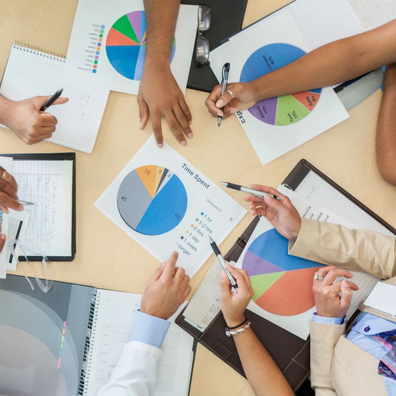 Pie charts and graphs are used to discuss hospital's financial situation. A group of doctors, nurses and hospital executives are gathered around a round board table. Photo taken from high angle viewpoint.