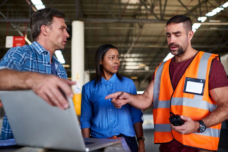 Shot of a group of coworkers talking together over a laptop while standing in a large warehouse