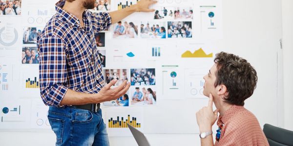 Two men discussing charts and graphs on a wall.