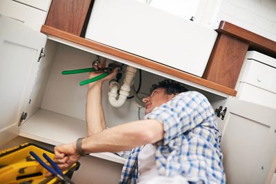Man fixing plumbing under a kitchen sink with a wrench. emergency plumbing, plumbing installation, plumbing repair 