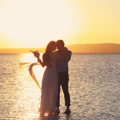 Couple embracing near water at sunset, bride holding bouquet.