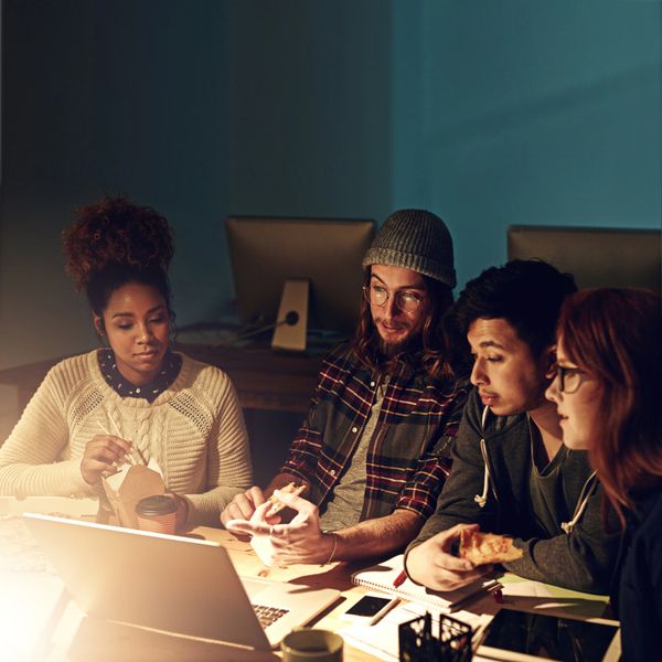 Four young adults eating and working together late at night around a laptop.