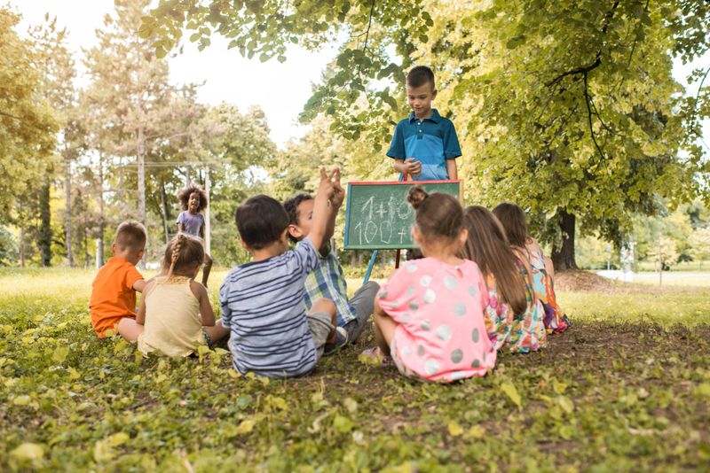 Little boy having fun with his friends in the park while explaining them mathematics on blackboard.
