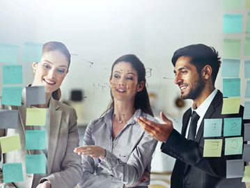 Three professionals collaborating and discussing ideas using sticky notes on a glass board.