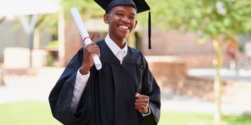 Smiling graduate in cap and gown holding diploma outdoors.