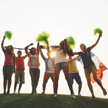 Group of young people cheering with green pom-poms and a flag at sunset.