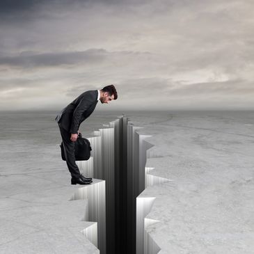 Businessman cautiously looks into a deep crack in the ground under a cloudy sky.