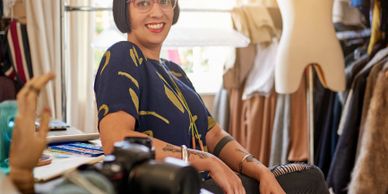 Smiling woman with glasses in a creative studio surrounded by fabrics and tools.