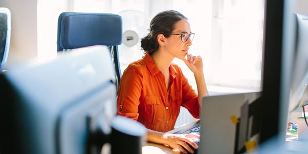 Woman in orange shirt working focused at a computer in a bright office.