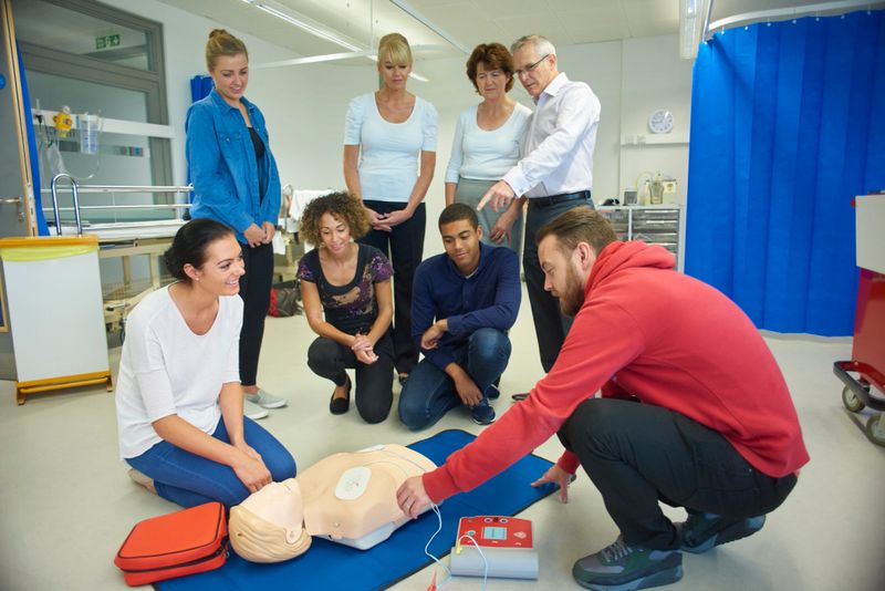 a mixed age group listen to their tutor as he shows the procedure involved to resuscitate using a defibrillator .