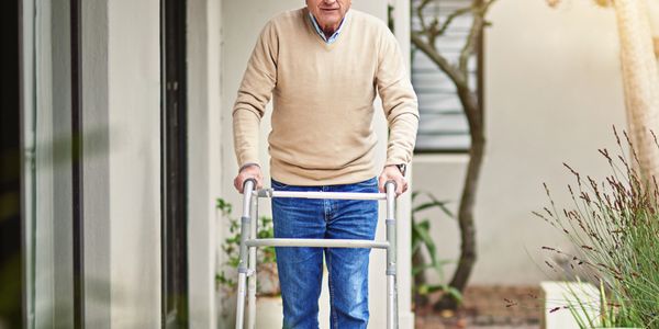 Older man using a walker outside a house on a sunny day.