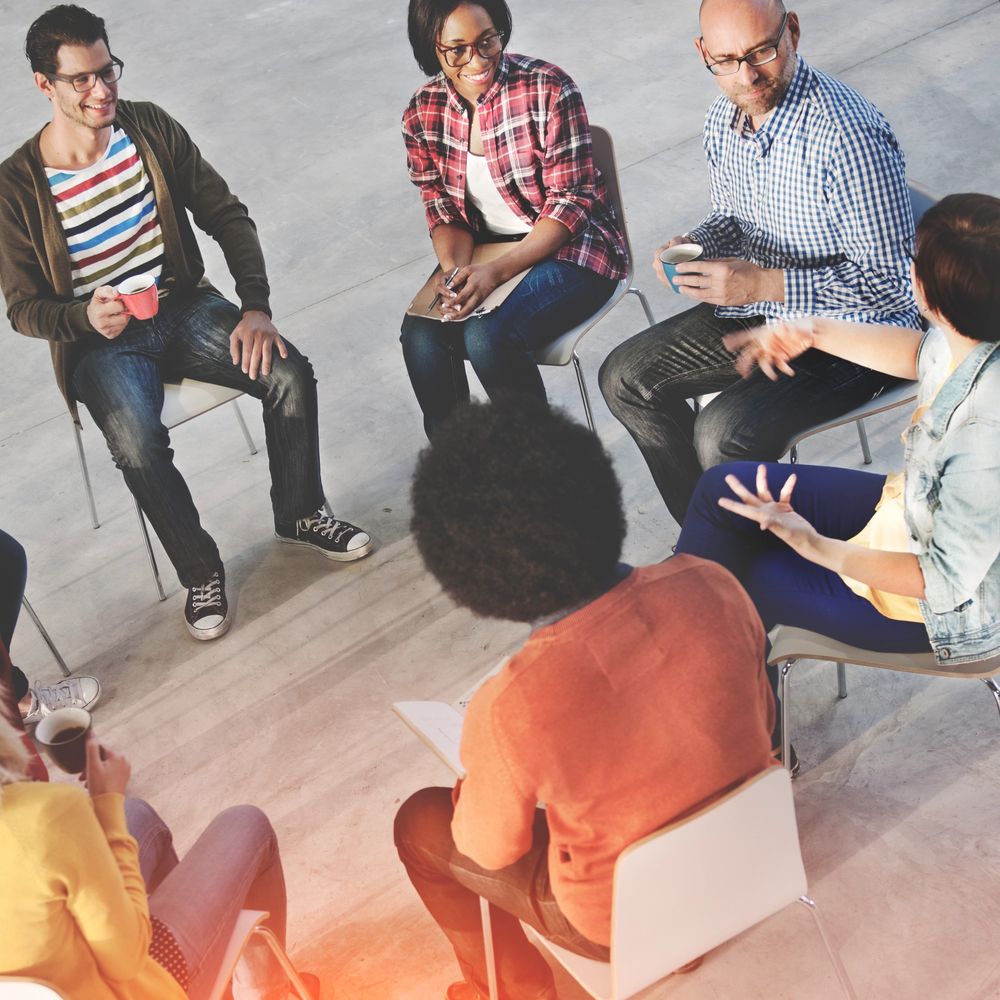 Diverse group in casual meeting, some holding coffee, engaging in discussion.