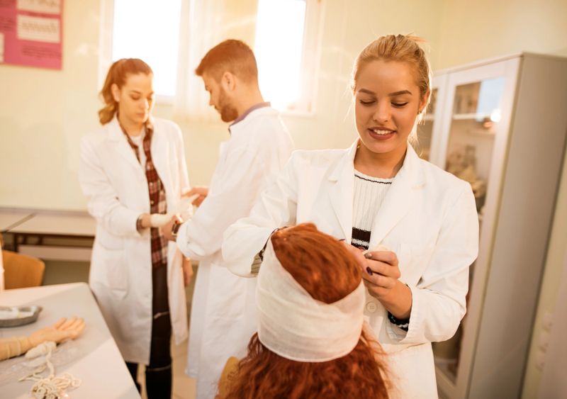 Group of medical students applying bandages on each other during first aid training course in laboratory. Focus is on smiling female student applying bandage on classmate's head.
