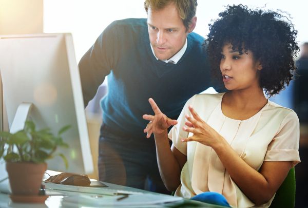 Two coworkers discuss work in front of a computer in a bright office.