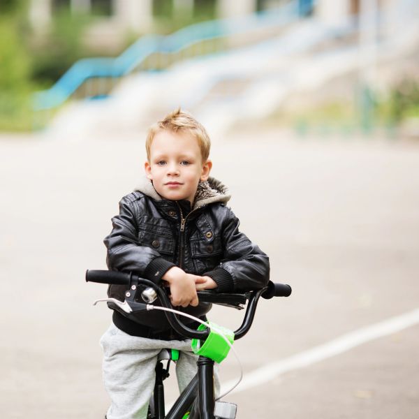 A young boy in a black jacket sitting on a bicycle outdoors.