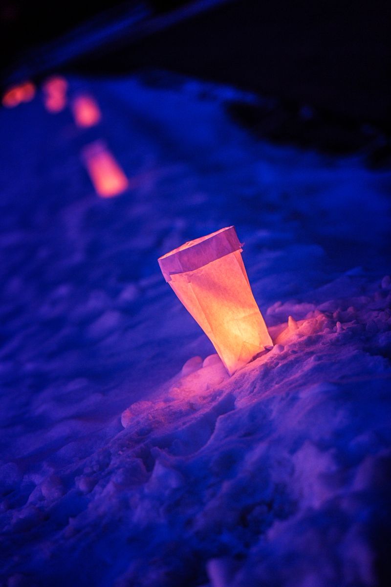 A Christmas Eve night line of symbolic candle light illuminated paper lantern paper bag luminaries arranged in a long row at the edge of a suburban neighborhood street. This type of decorative light luminary/luminaria/farolitos is assembled with sand and a small wax votive candle in lunch-size paper bags. Every year, many families in this Rochester, New York, northeastern USA area residential housing development cooperate by putting luminaries along the perimeter of their property at the edge of the street - creating a coordinated, symbolic and tranquil community holiday event to emphasize togetherness and "Peace on Earth" throughout the neighborhood.