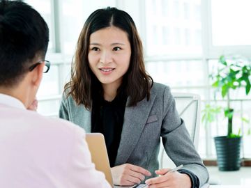A woman in a gray blazer converses with a man in an office setting.
